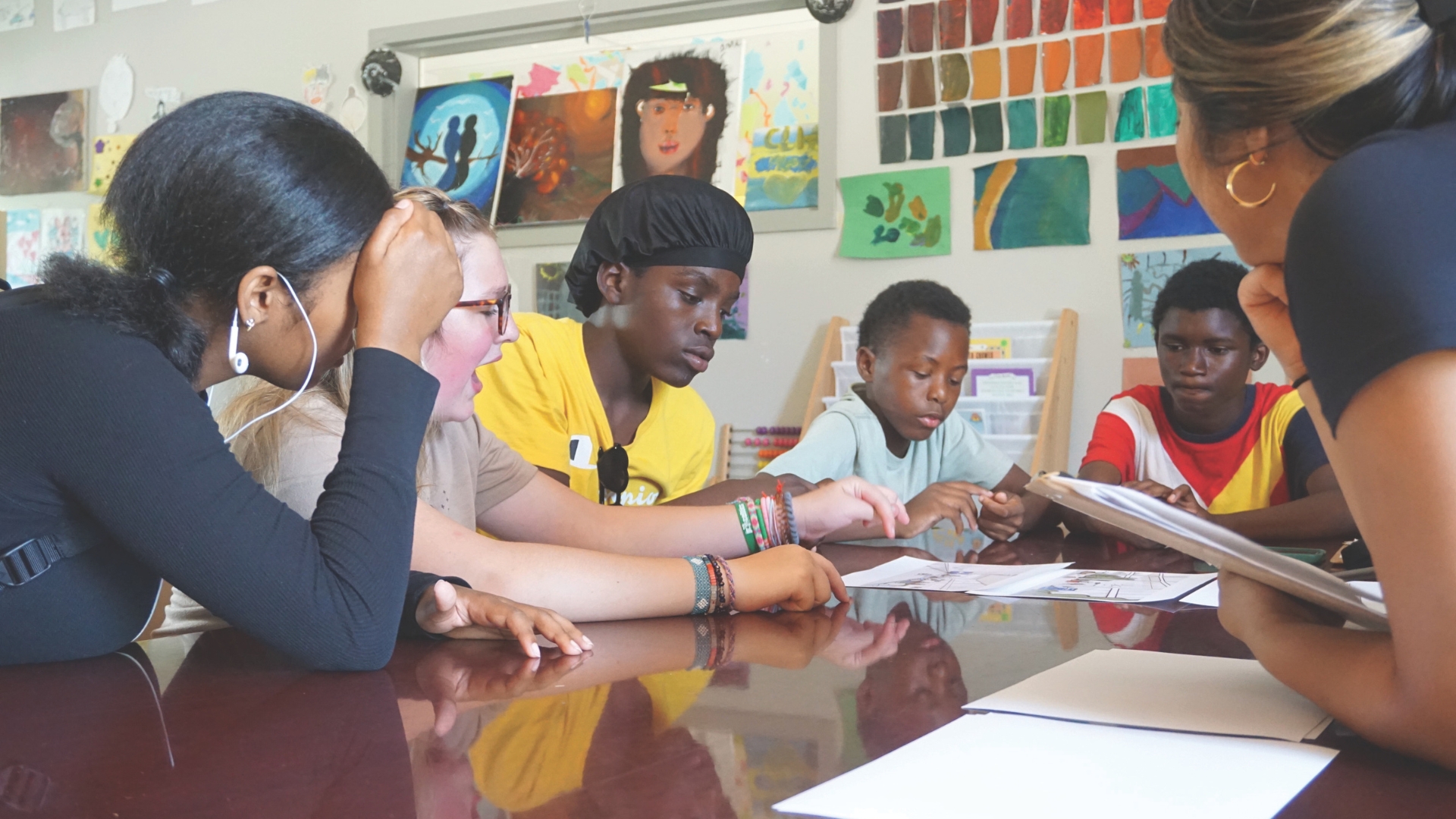 A group of diverse young people sit around a table, focusing on papers and worksheets in front of them. The room is decorated with colorful artwork and posters on the walls, creating a creative and educational atmosphere. The children are engaged, leaning in toward each other in discussion.