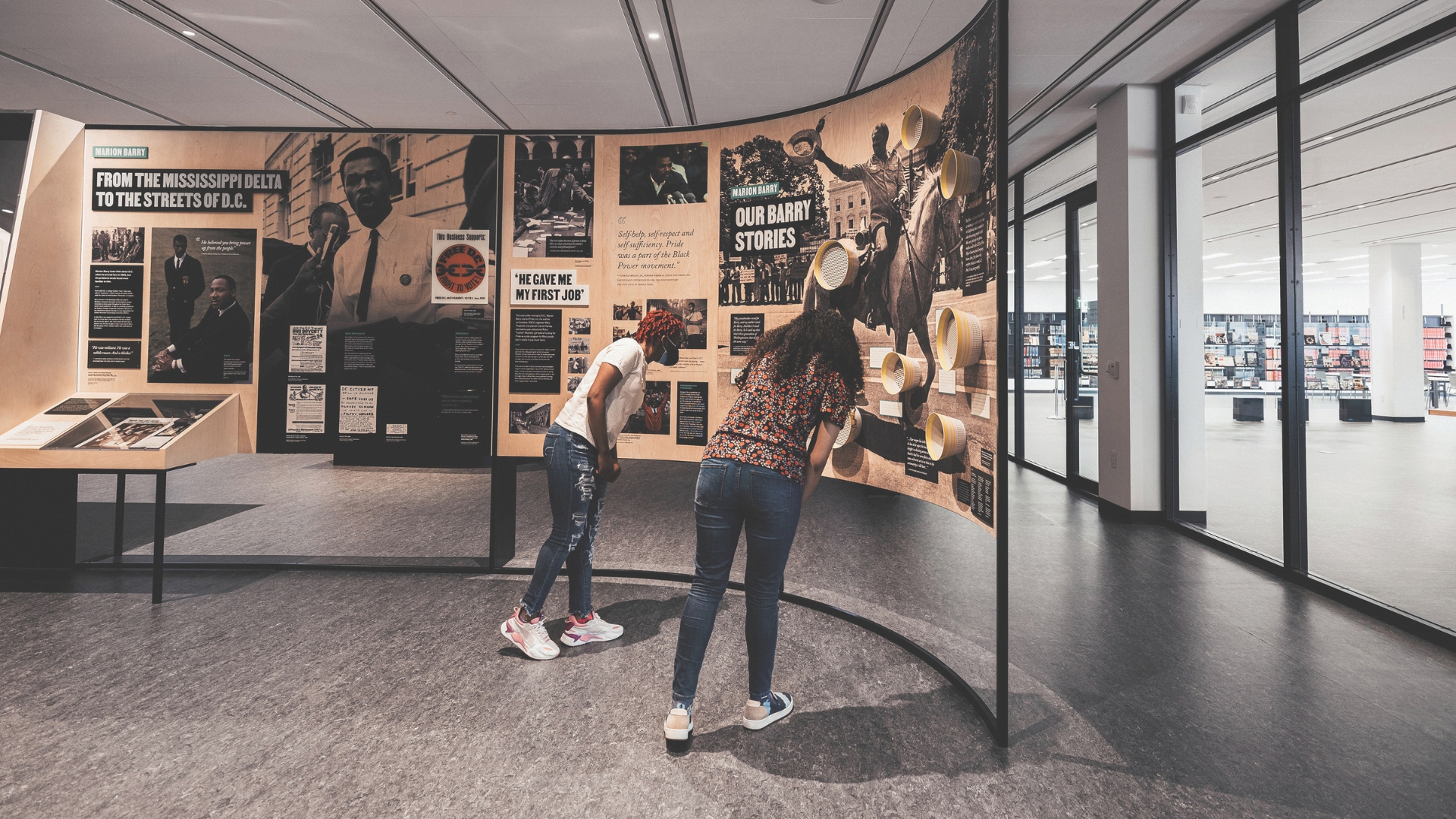 Two people, a man and a woman, lean forward closely examining a curved exhibit panel that features images and text about the life of Marion Barry. The exhibit is located in a modern, open space with large windows in the background. The man on the left is wearing casual jeans and a white t-shirt, while the woman on the right is wearing a patterned shirt and jeans.