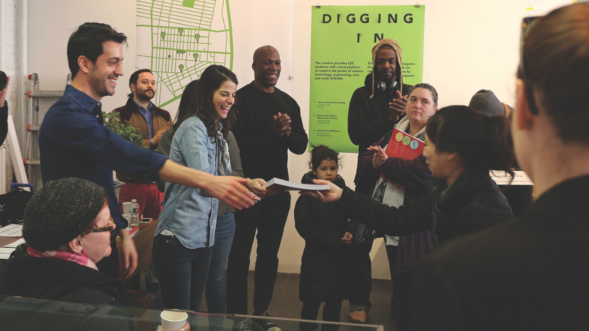 A group of people are gathered in a lively room, celebrating an event. One man is handing over a piece of paper to another person, with others smiling and clapping in the background. The space has posters and informational graphics on the wall, one prominently labeled "DIGGING IN," and the atmosphere is casual and collaborative.