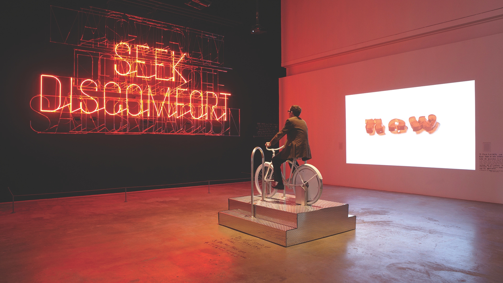 A person sits on a stationary white bicycle in a dimly lit room. In the background, a neon sign in bright red letters reads, "SEEK DISCOMFORT." A projection on the wall displays the word "now" in bold text. The atmosphere is interactive and thought-provoking.