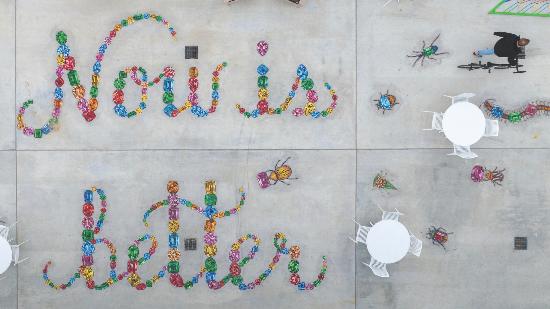 An overhead view of an outdoor space featuring colorful text and insect-like sculptures. The text spells out “Now is Better” in bright, jewel-like colors, surrounded by oversized beetle and bug sculptures. White tables and chairs are scattered throughout the space, and a person with a bicycle walks along the perimeter.