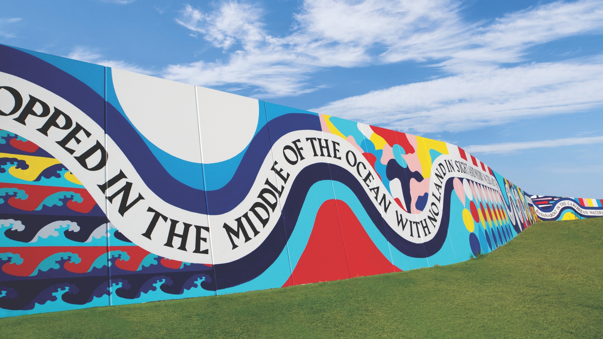 A vibrant mural painted on a long outdoor wall under a blue sky. The mural features colorful abstract shapes and waves, with bold black text that reads, "DROPPED IN THE MIDDLE OF THE OCEAN WITH NO LAND IN SIGHT," flowing across the design.