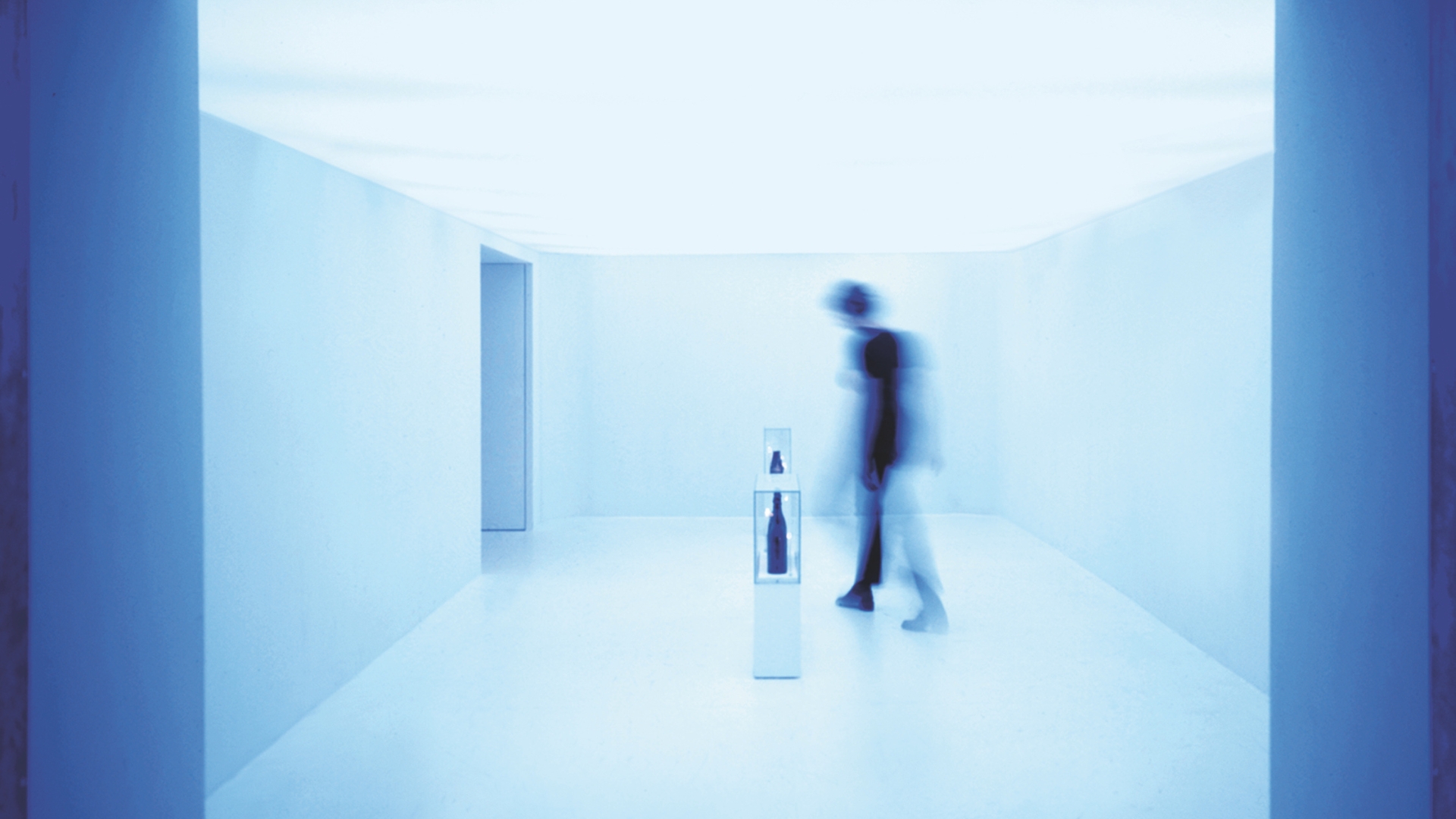 A person walks through a minimalistic, all-blue room, passing two small pedestal displays with objects encased in glass. The room is softly lit, and the person is captured in motion, creating a blurred effect.