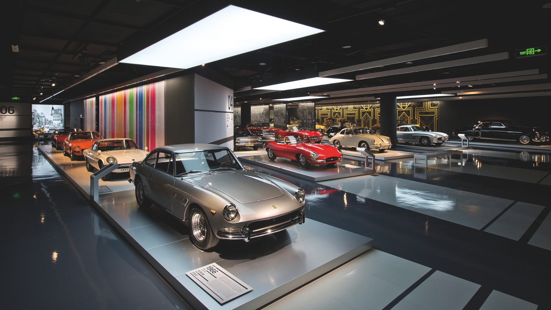 A sleek, modern car exhibit featuring a row of vintage cars displayed on platforms. The space is well-lit, with colorful vertical stripes on the wall and various luxury vehicles, including a silver car in the foreground and red cars further back.