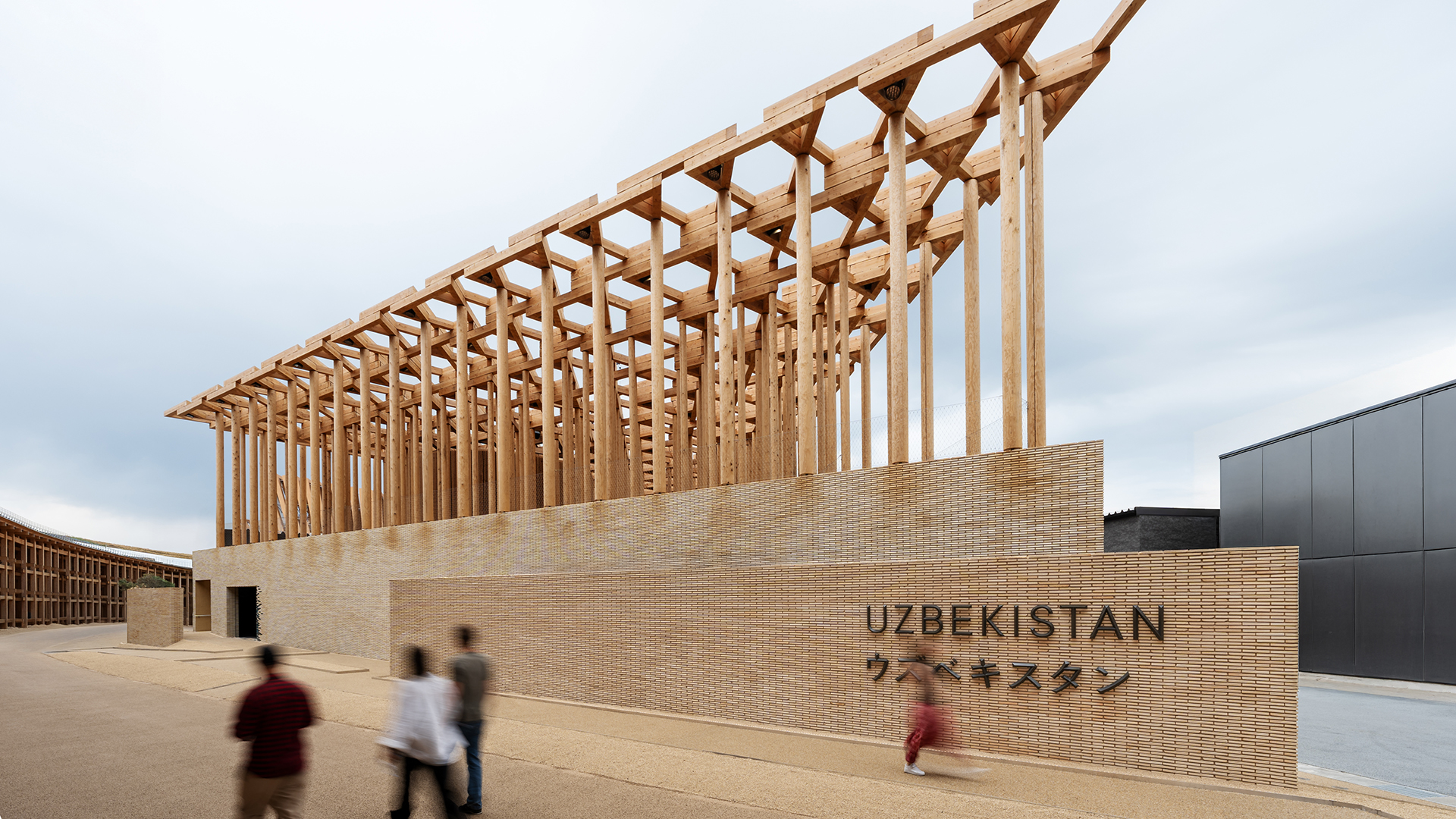 Exterior of the Uzbekistan Pavilion showing timber colonnade above a brick base with visitors walking past.