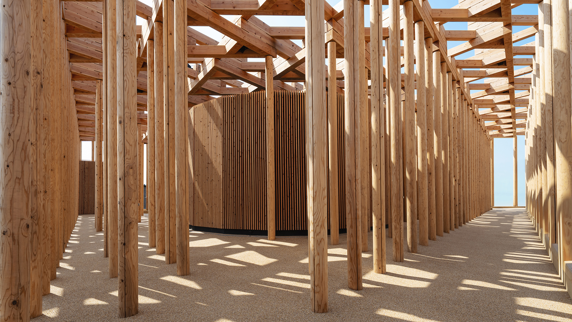 Interior view of the wooden terrace with dense vertical columns casting shadows on the ground.