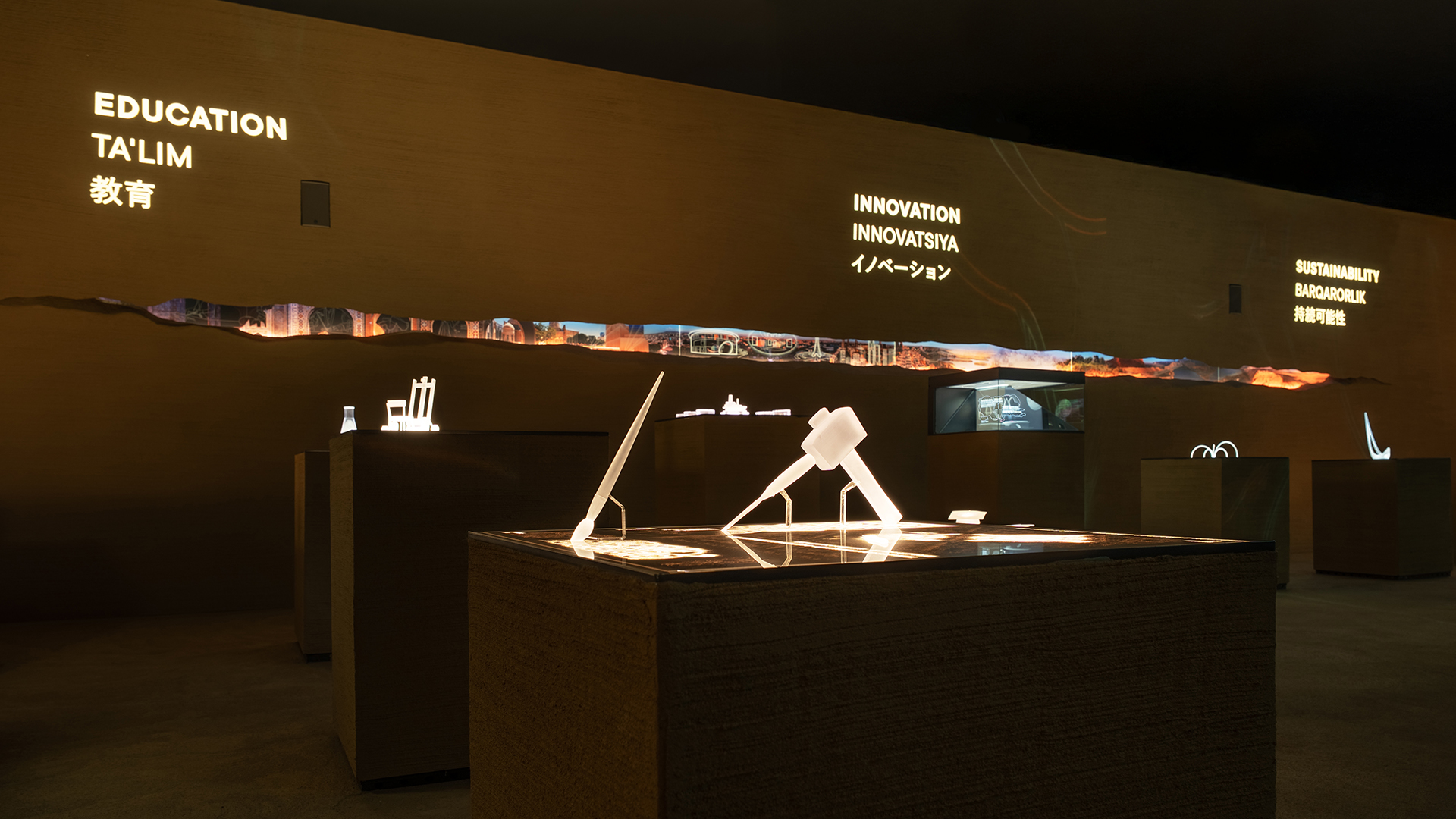 Dark exhibition space featuring glowing models and wall projections labeled Education, Innovation, and Sustainability.