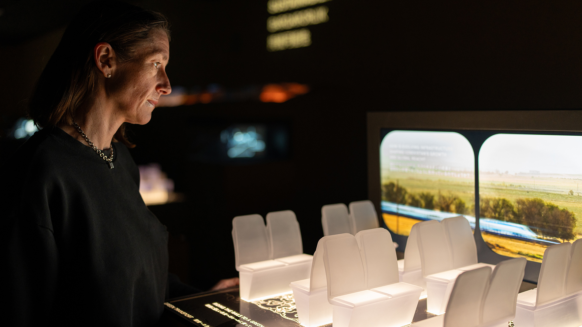 A woman observes an interactive model of train seats with a projected window showing Uzbek countryside.