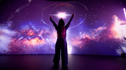 A visitor raises their arms toward a galaxy projection in the Colors of Creation immersive experience at The Weitzman.